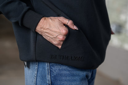 A close-up of a person wearing a black hoodie with a hand tucked into the front pocket. The hoodie features the text 'BE THE LIGHT' debossed on the lower pocket.