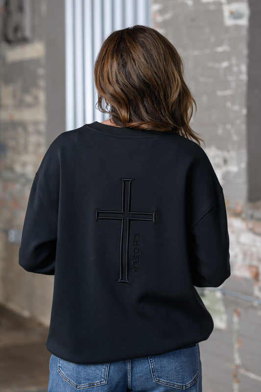 A person viewed from behind wearing a black crewneck sweatshirt with a large tonal cross and vertical 'Chosen' text on the back, paired with blue jeans in an urban setting.