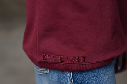 Close-up of a person wearing a burgundy crewneck sweatshirt with 'BE THE LIGHT' embroidered on the hem, paired with blue jeans.