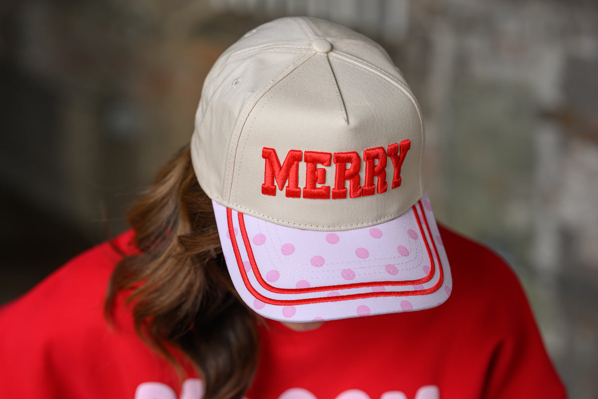 A person wearing a beige baseball cap with 'MERRY' embroidered in red on the front and a pink polka-dot brim with red piping, paired with a red sweatshirt against a blurred gray background.