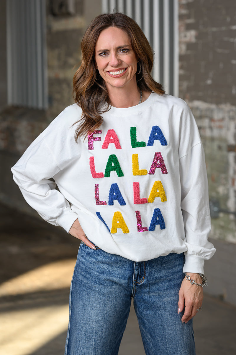 A woman wearing a white crewneck sweatshirt with 'FA LA LA LA LA LA' printed in colorful glitter-textured letters, paired with blue jeans, standing in front of a rustic brick wall.