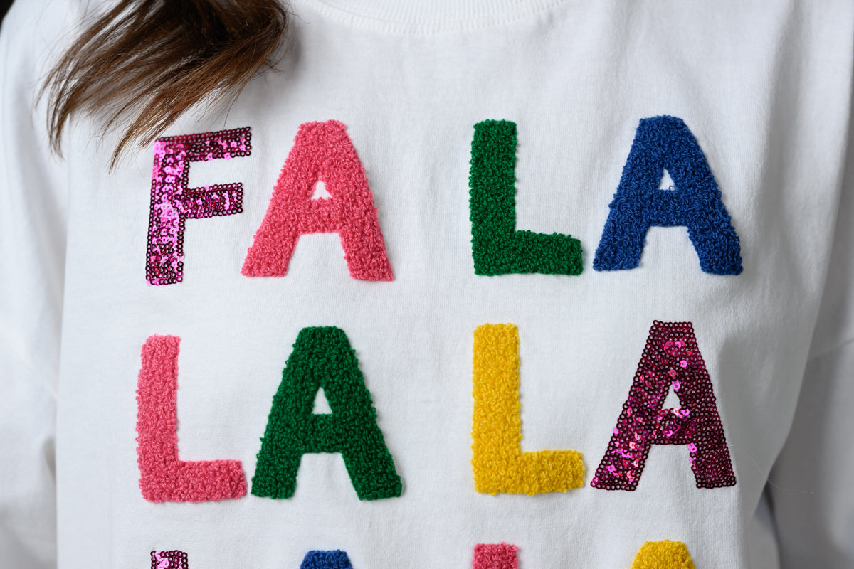 A close-up of a white crewneck sweater featuring colorful, textured sequin letters spelling out 'FA LA LA LA' 