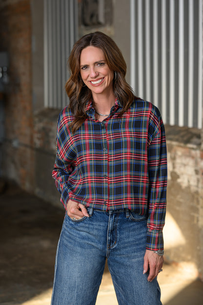 A woman smiling and wearing a red, navy, and blue plaid button-down shirt with blue jeans, standing in front of a rustic brick and corrugated metal background.