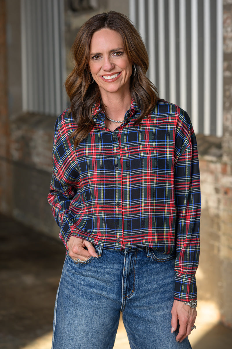 A smiling woman with brown hair stands wearing a long-sleeve button-up plaid shirt in navy, red, and white, paired with blue jeans, in front of a rustic brick and corrugated metal background.