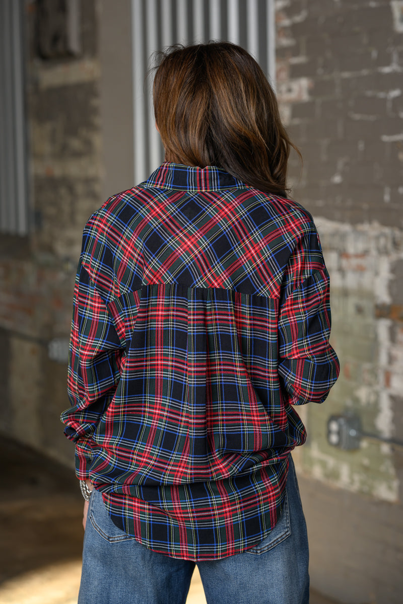 A woman viewed from behind wearing a red, navy, and blue plaid button-up shirt and blue jeans, standing in front of a rustic brick wall.