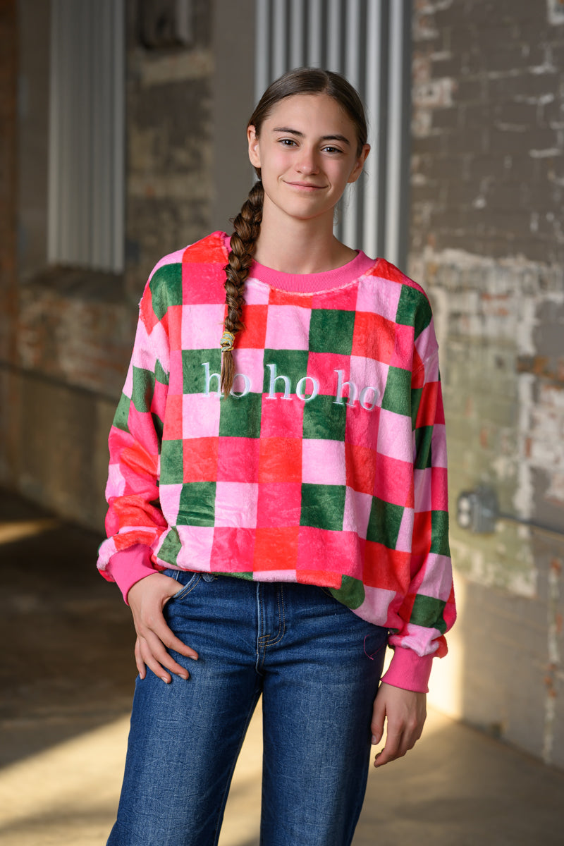 A young woman with a long braid stands in a rustic indoor setting wearing a pink, red, and green checkered sweatshirt with 'ho ho ho' printed on the front, paired with blue jeans.
