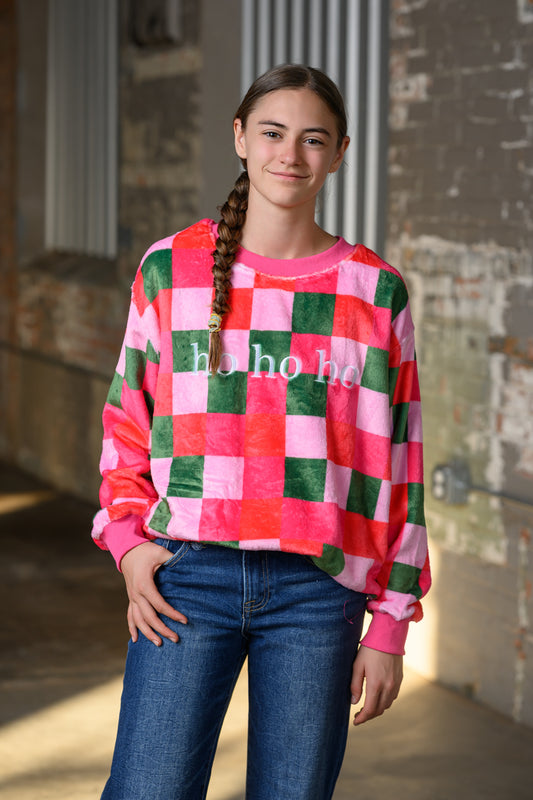 A young woman with a long braid stands in a rustic indoor setting wearing a pink, red, and green checkered sweatshirt with 'ho ho ho' printed on the front, paired with blue jeans.