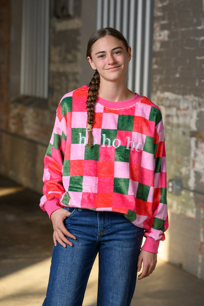 A young woman with a long braid stands in an industrial alleyway wearing a pink, green, and white checkered sweatshirt with 'h ho h ho' printed in white across the chest, paired with blue jeans.