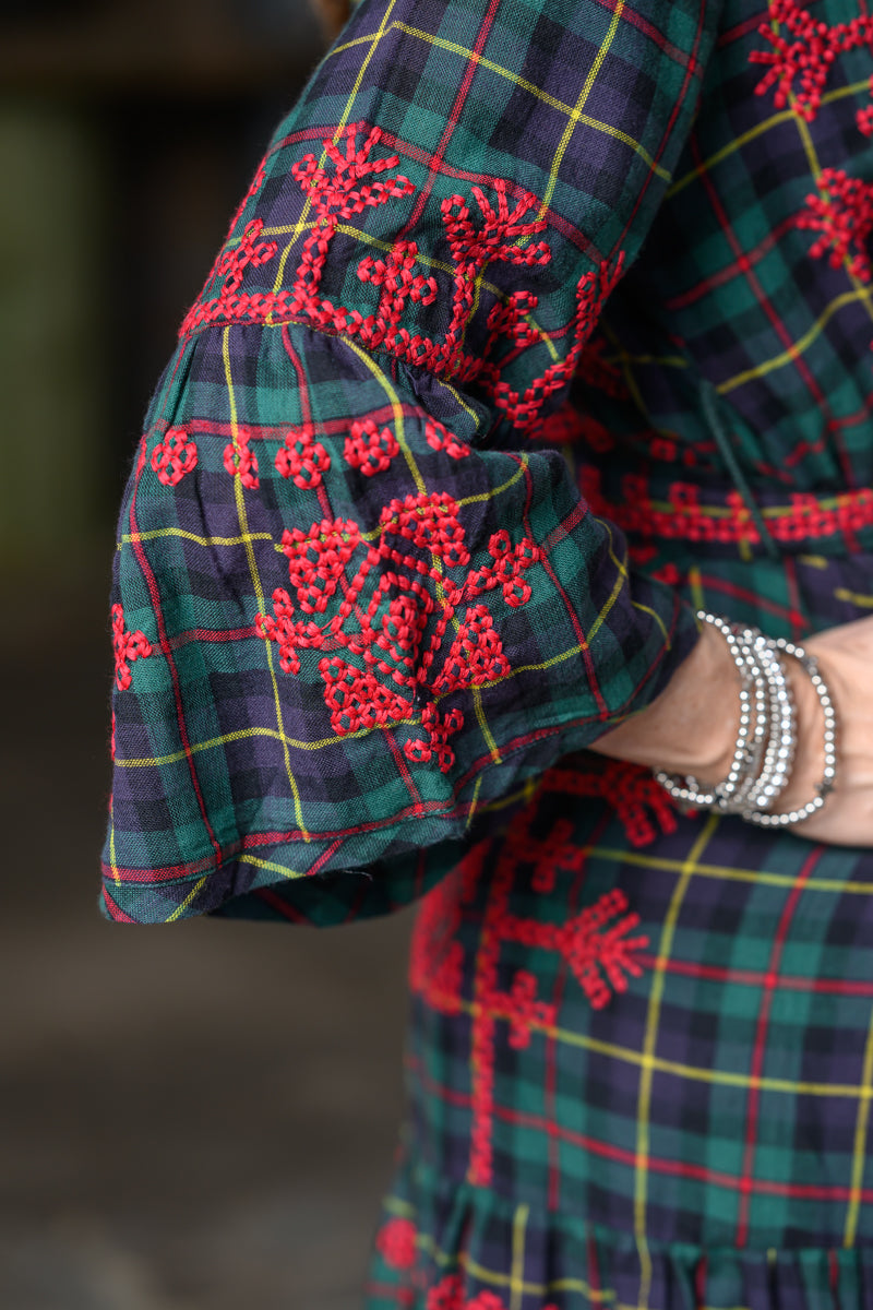 Close-up of a person wearing a green tartan plaid garment with red embroidered floral patterns and silver beaded bracelets on the wrist.