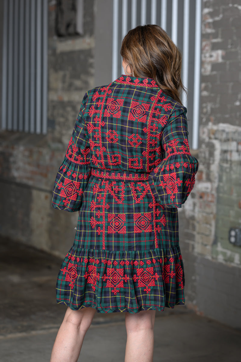 A woman viewed from behind wearing a green tartan plaid coat with red embroidered geometric patterns, set against a rustic brick and concrete wall background.