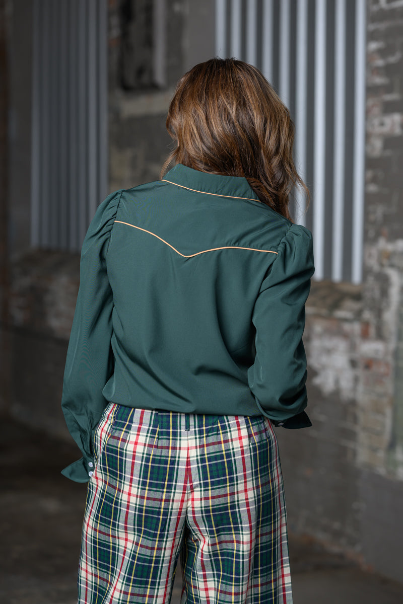 A woman viewed from behind wearing a dark green western-style long-sleeve shirt with contrast stitching and green plaid pants in an industrial alleyway.