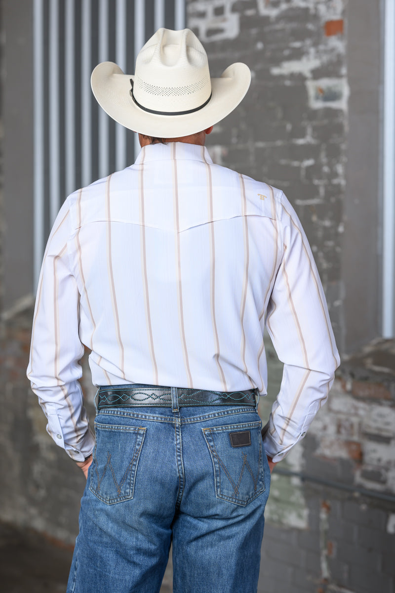 A man viewed from behind wearing a white western-style long-sleeve shirt with thin tan vertical pinstripes, a cream cowboy hat, and blue jeans, standing against a gray brick wall.
