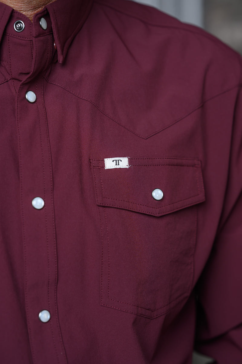 Close-up of a burgundy western-style button-down shirt with a chest pocket and a small white label featuring a monogram logo.