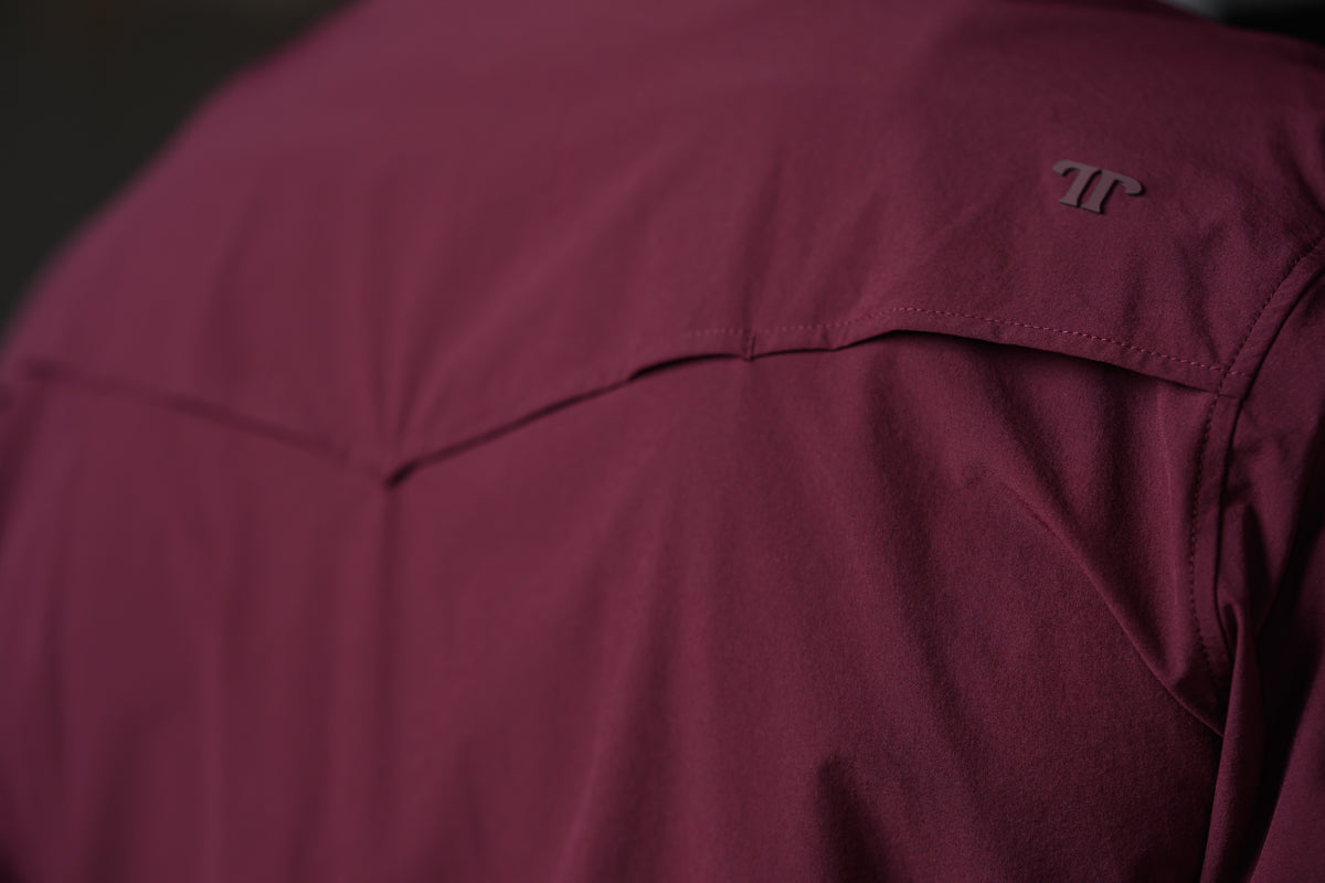 Close-up of a burgundy technical outdoor jacket showing the shoulder seam, stitching, and a dark embroidered logo on the upper sleeve.