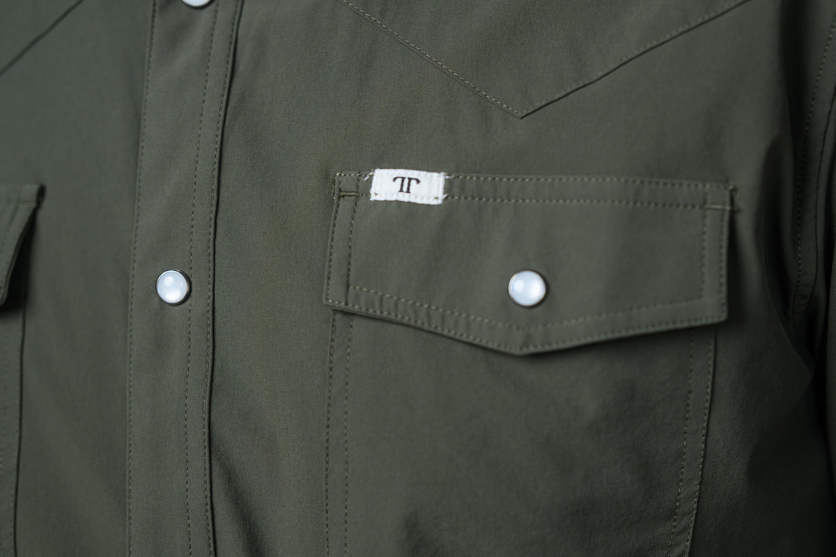 Close-up of an olive green western-style shirt jacket featuring a chest pocket with a white snap button and a small white brand label with a black monogram.
