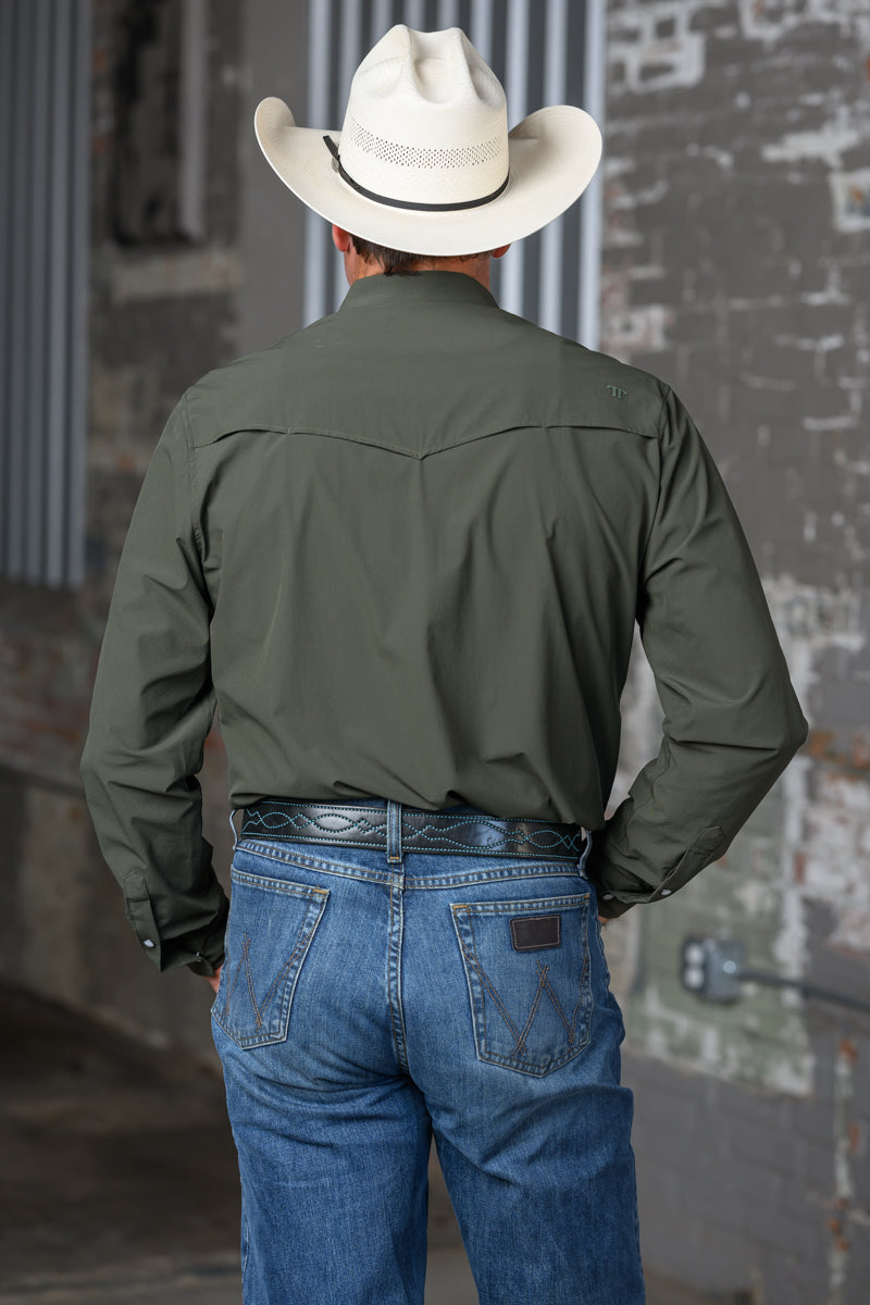 A man viewed from behind wearing an olive green long-sleeve western shirt, blue jeans, a black western belt, and a white cowboy hat, standing against a rustic brick wall.