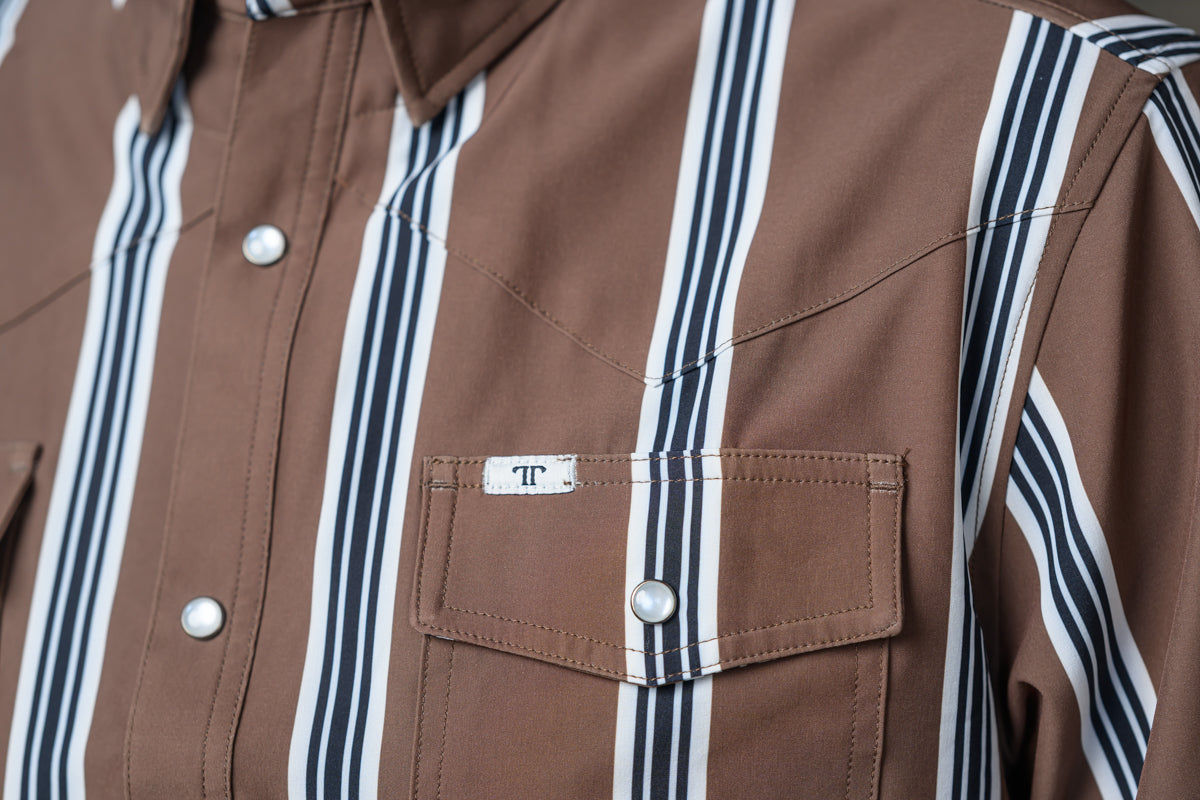 Close-up of a brown western-style shirt with vertical black and white stripes, featuring a chest pocket with a small white label that has a black monogram logo.