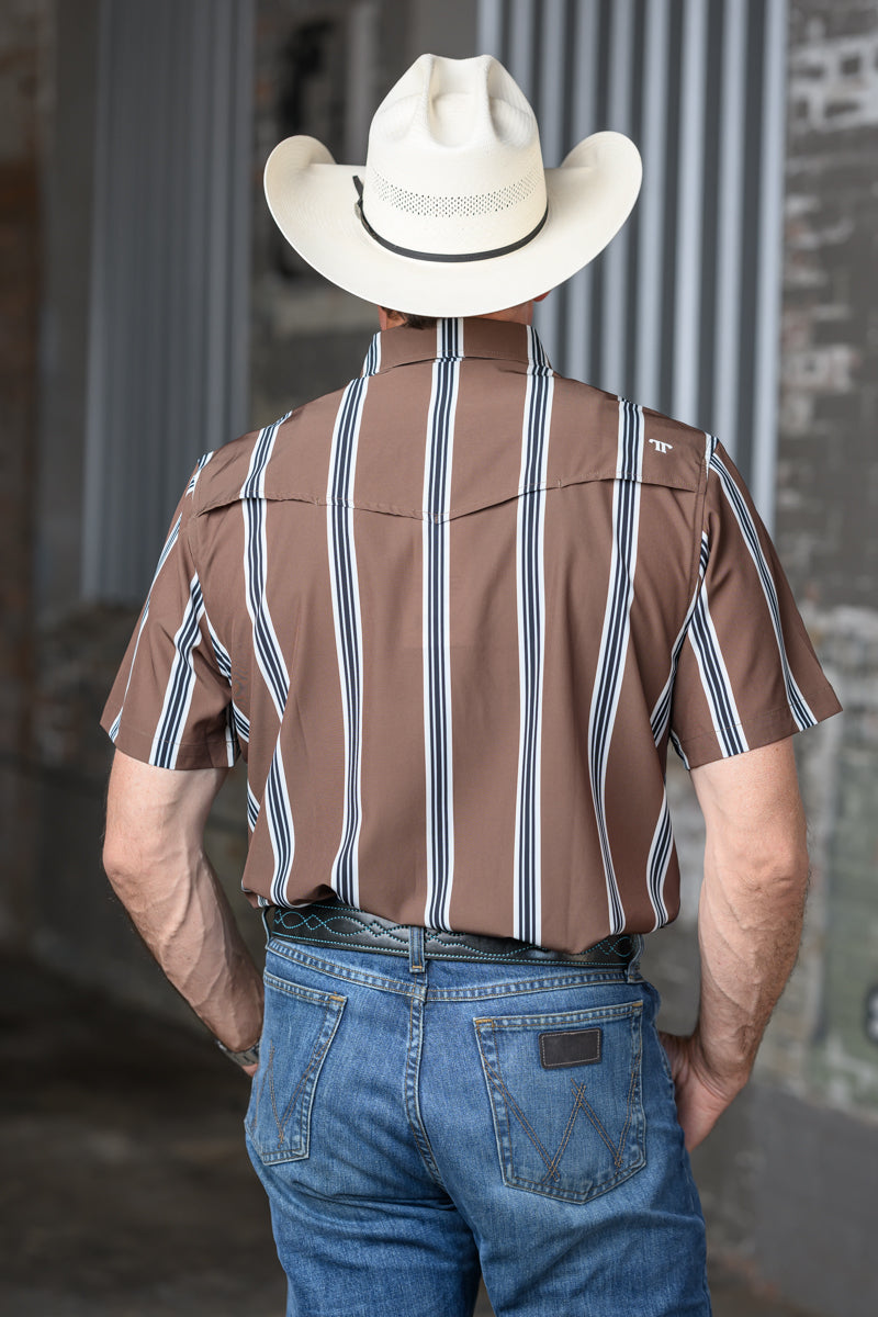 A man viewed from behind wearing a brown short-sleeve button-up shirt with vertical white and navy stripes, blue jeans, and a white cowboy hat in front of a corrugated metal wall.
