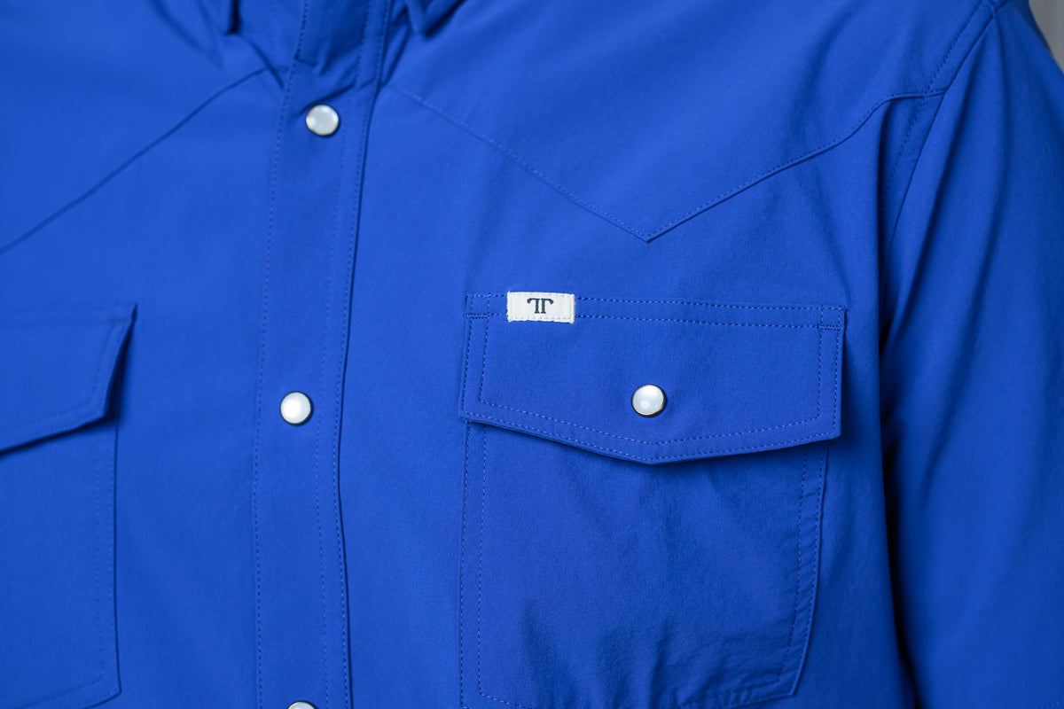 A close-up of a bright royal blue long-sleeve western-style shirt with white snap buttons and a chest pocket featuring a small white label with a black 'FF' logo.