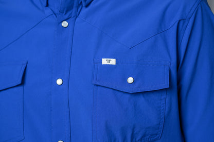 A close-up of a bright royal blue long-sleeve western-style shirt with white snap buttons and a chest pocket featuring a small white label with a black 'FF' logo.