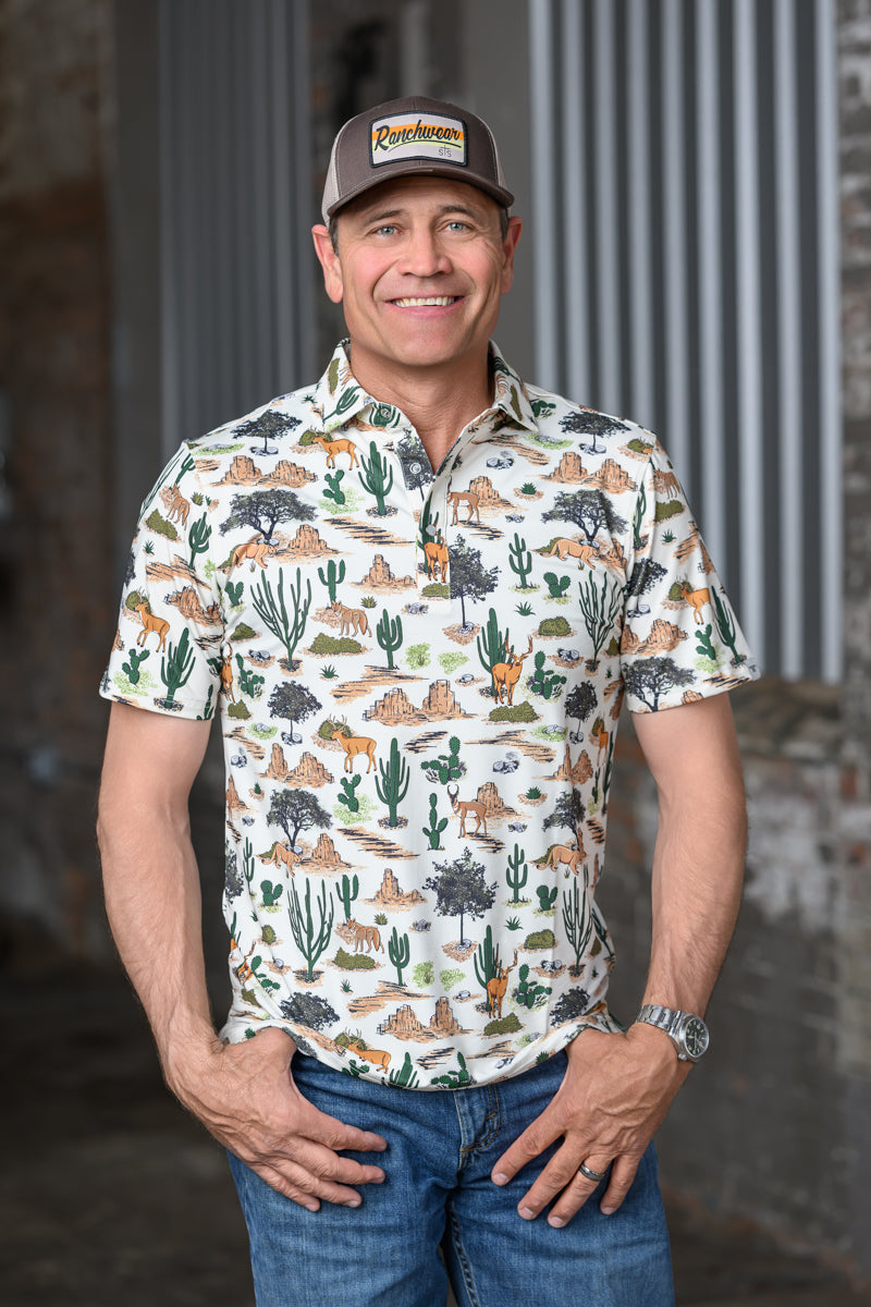 A smiling man wearing a short-sleeve button-up shirt with a desert animal and cactus print, blue jeans, and a brown baseball cap with a visible 'Cowhide' logo patch, standing against a rustic gray and brown background.
