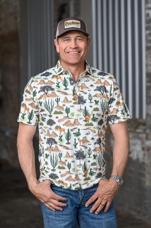 A smiling man wearing a short-sleeve button-up shirt with a desert animal and cactus print, blue jeans, and a brown baseball cap with a visible 'Cowhide' logo patch, standing against a rustic gray and brown background.