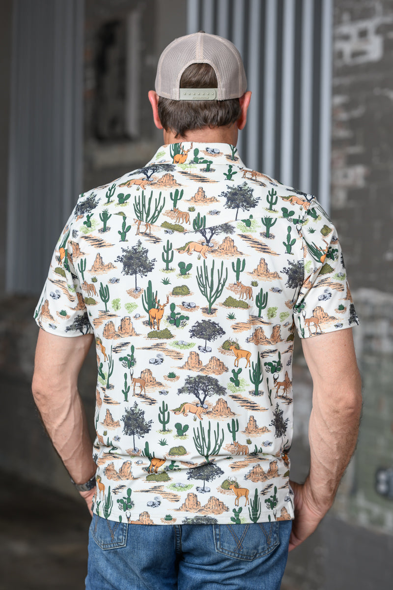 A man viewed from behind wearing a short-sleeve button-up shirt with a desert landscape print, featuring cacti, mountains, and vehicles on an off-white background. He is also wearing a beige baseball cap and blue jeans.