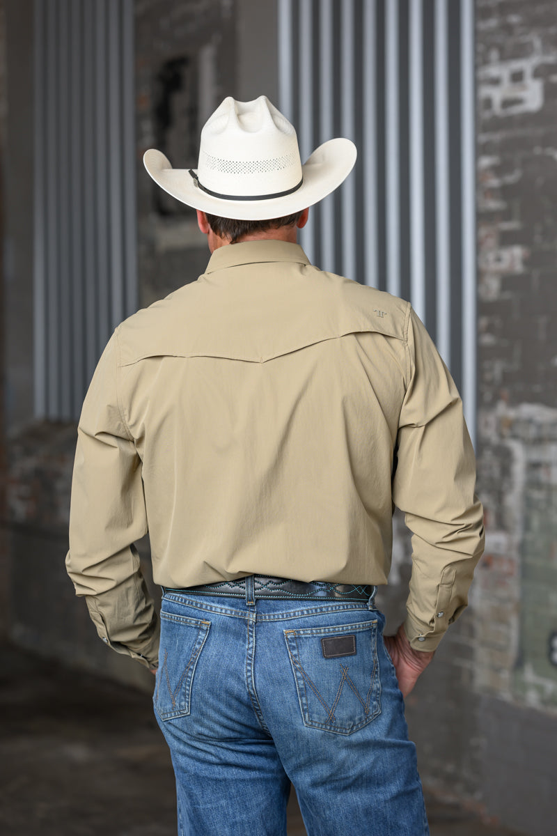 A man viewed from behind wearing a light beige western-style long-sleeve shirt, blue jeans, and a white cowboy hat, standing in front of a gray corrugated metal wall.