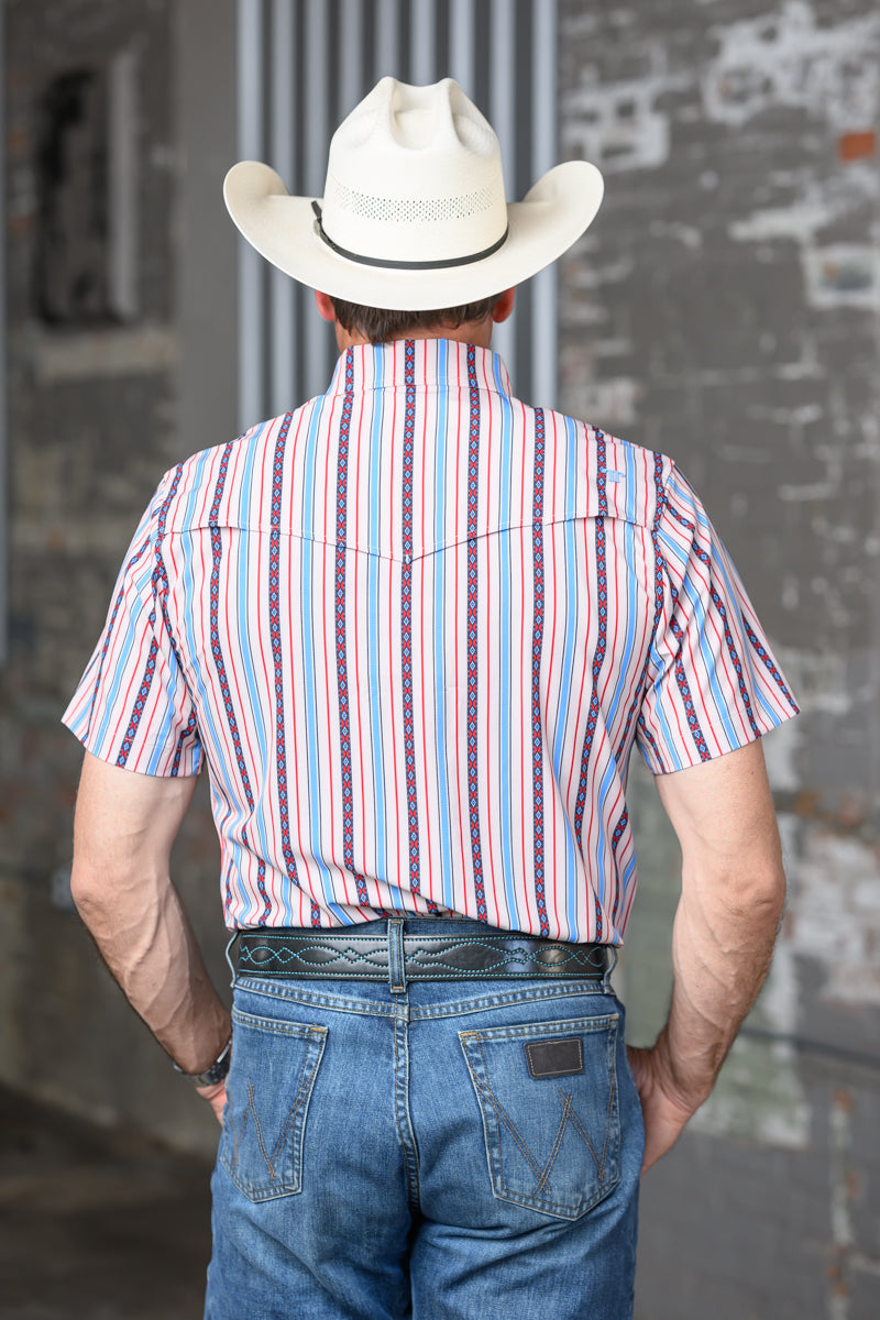 A man viewed from behind wearing a cream cowboy hat, a short-sleeve western shirt with vertical red, blue, and white stripes, and blue jeans, standing against a gray brick wall.