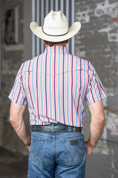 A man viewed from behind wearing a cream cowboy hat, a short-sleeve western shirt with vertical red, blue, and white stripes, and blue jeans, standing against a gray brick wall.