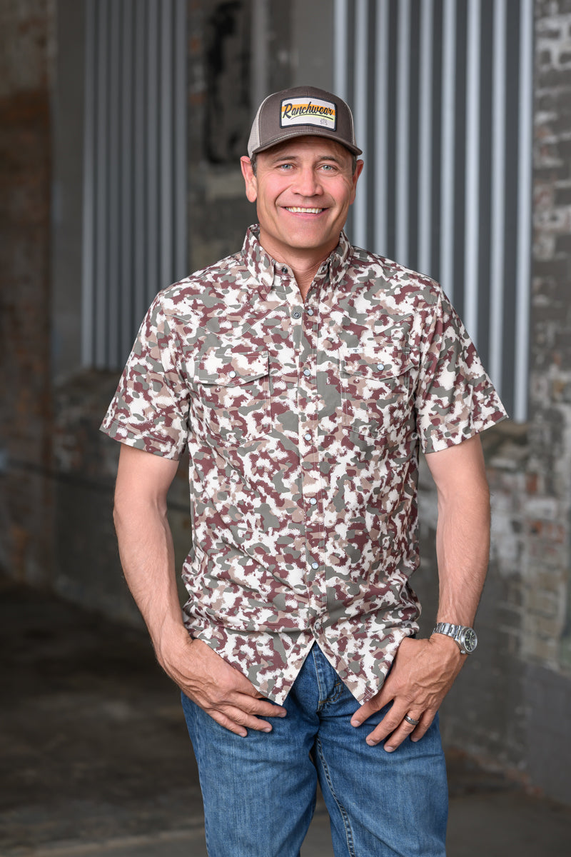 A man wearing a short-sleeve button-up shirt with a brown, beige, and white camouflage pattern, blue jeans, and a baseball cap, standing in front of a rustic brick and corrugated metal wall.