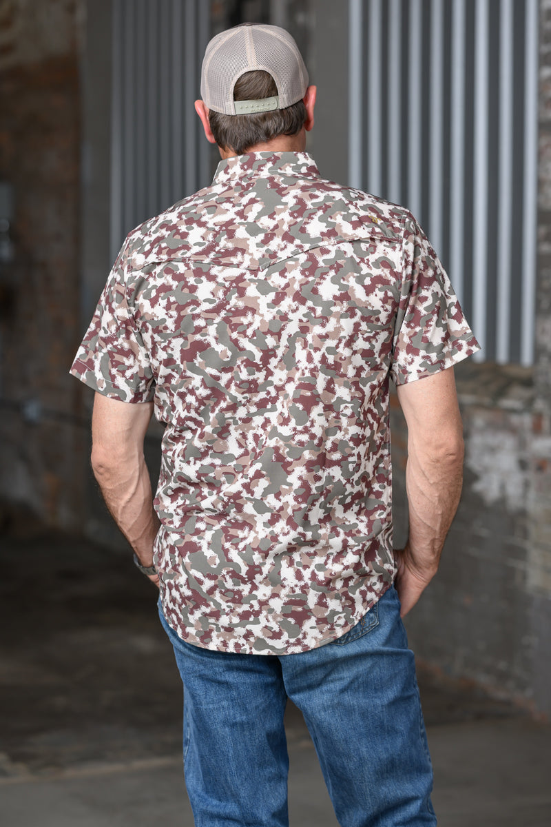 A man viewed from behind wearing a short-sleeve button-up shirt with a beige, brown, and gray camouflage pattern, paired with blue jeans and a beige baseball cap.