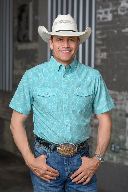 A man wearing a turquoise short-sleeve western button-up shirt with a subtle geometric pattern, blue jeans, a black belt, and a large decorative silver buckle, standing in front of a rustic gray brick wall.
