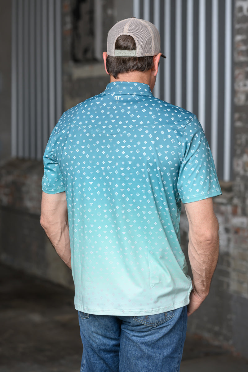 A man viewed from behind wearing a short-sleeve collared polo shirt with a white geometric pattern over a blue-to-light-teal gradient, paired with a beige baseball cap and blue jeans.