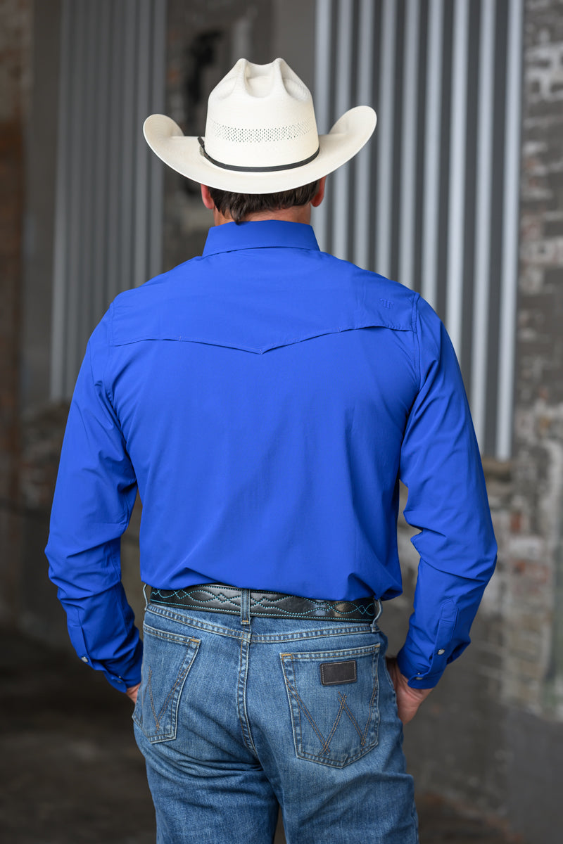 A man viewed from behind wearing a bright royal blue long-sleeve western shirt, a white cowboy hat, and blue jeans, standing in front of a corrugated metal and brick wall.