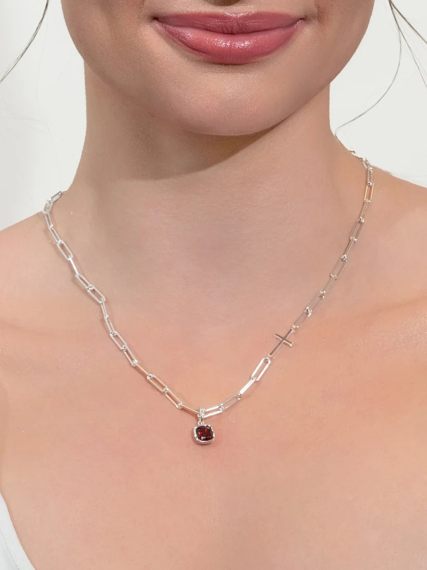 A close-up of a model's neck and chest wearing a silver paperclip chain necklace with a small round red gemstone pendant.