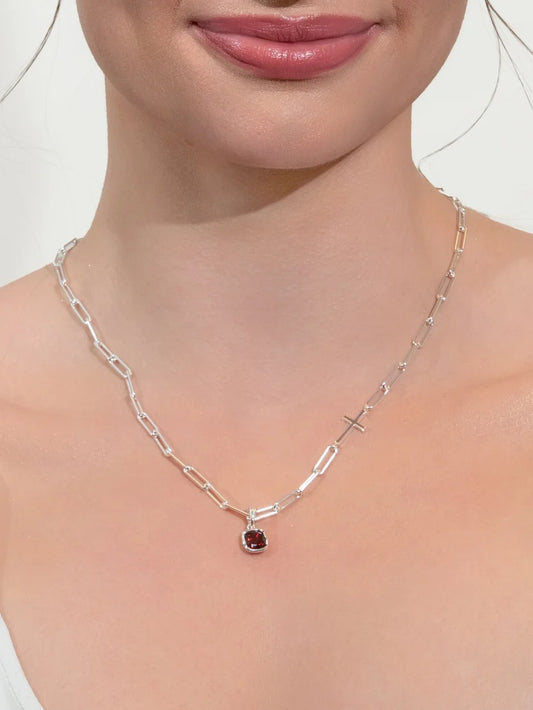 A close-up of a model's neck and chest wearing a silver paperclip chain necklace with a small round red gemstone pendant.