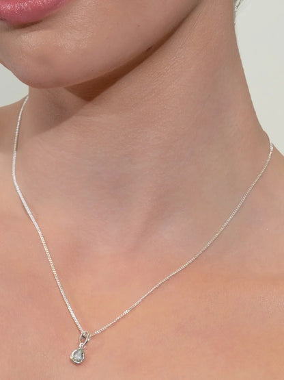 A close-up of a model's neck and lower face wearing a delicate silver chain necklace with a small teardrop-shaped clear gemstone pendant.