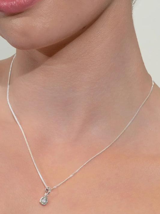 A close-up of a model's neck and lower face wearing a delicate silver chain necklace with a small teardrop-shaped clear gemstone pendant.