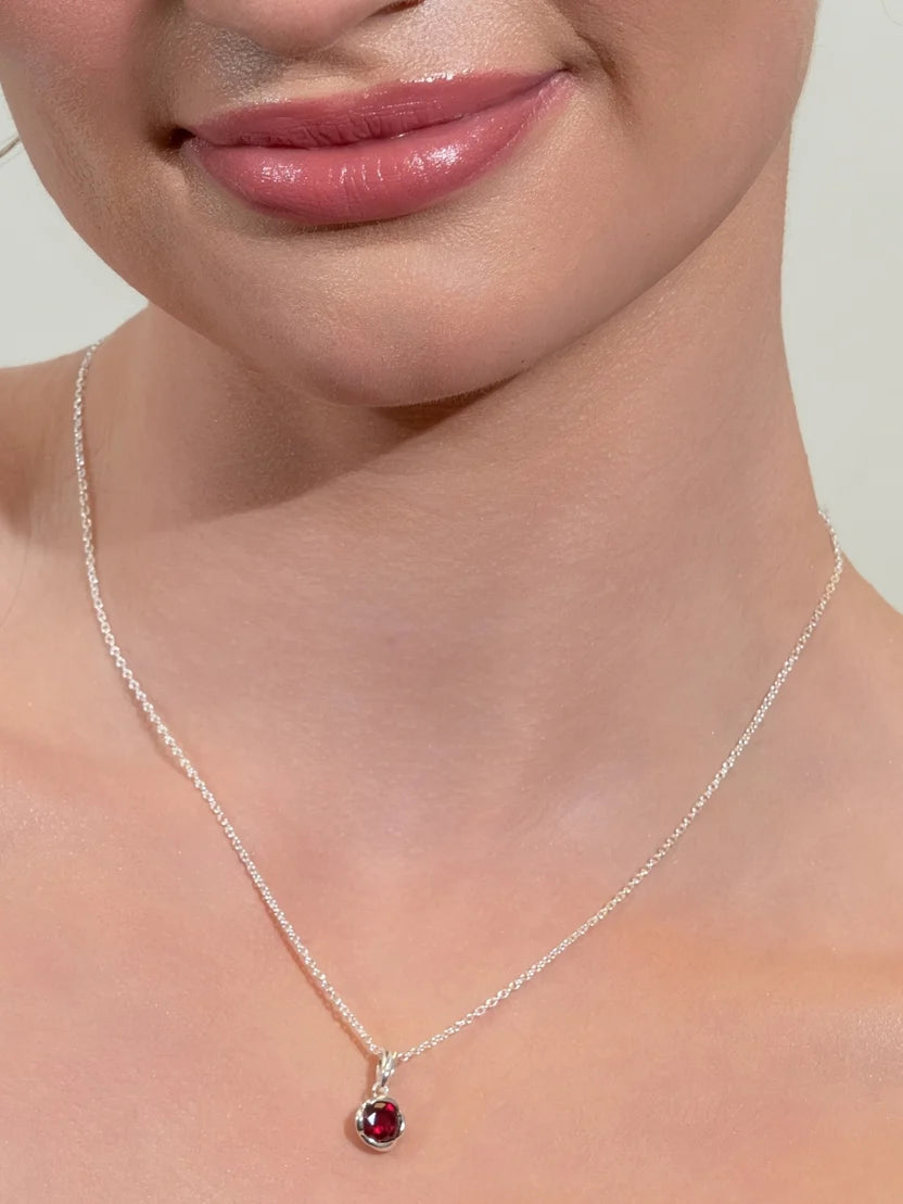 A close-up of a model's neck and lower face wearing a delicate silver chain necklace with a small oval red gemstone pendant.