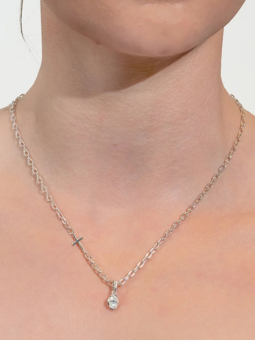 A close-up of a model's neck and upper chest wearing a delicate silver chain necklace with a small cross charm and a clear teardrop-shaped crystal pendant.