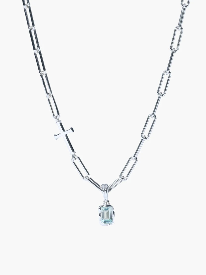 A silver paperclip chain necklace featuring a small silver cross pendant and a central bezel-set light blue gemstone, shown against a plain white background.