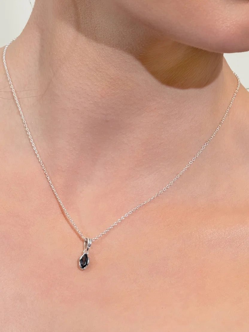 A close-up of a delicate silver chain necklace with a small teardrop-shaped dark gemstone pendant, worn on a model's neck.