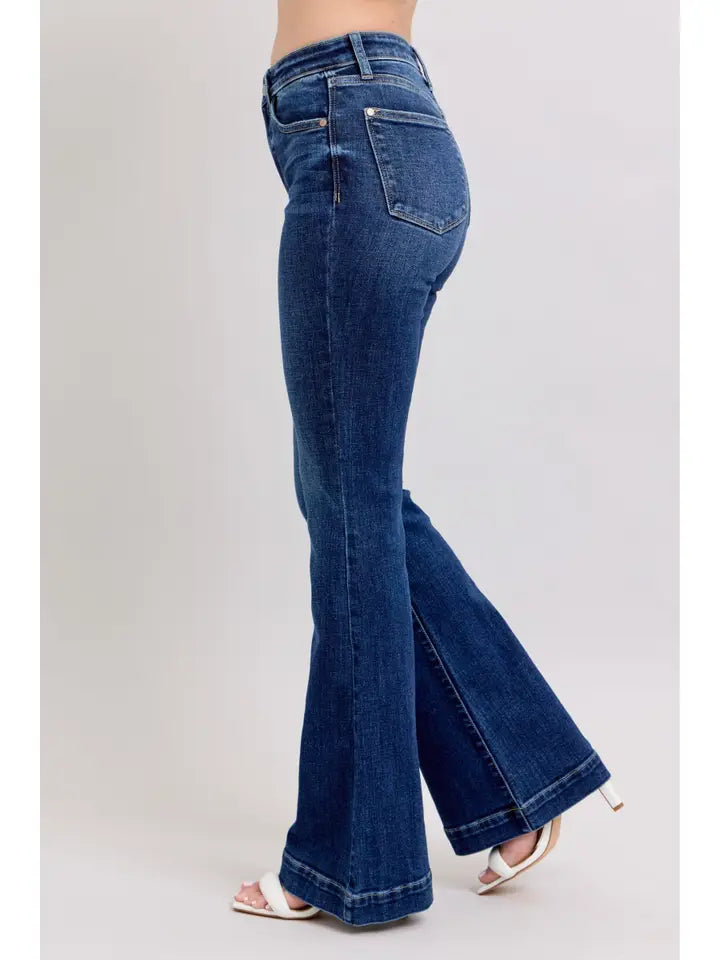 A person wearing dark blue flare jeans with a side-back view, paired with white heeled sandals, against a plain light gray studio background.