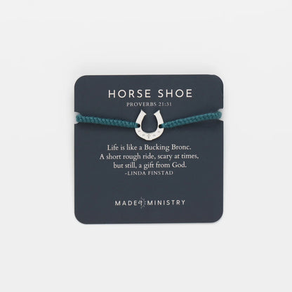 A silver horse shoe charm bracelet on a dark teal cord, presented on a dark gray card with a Bible verse quote and the brand name Made by Ministry.