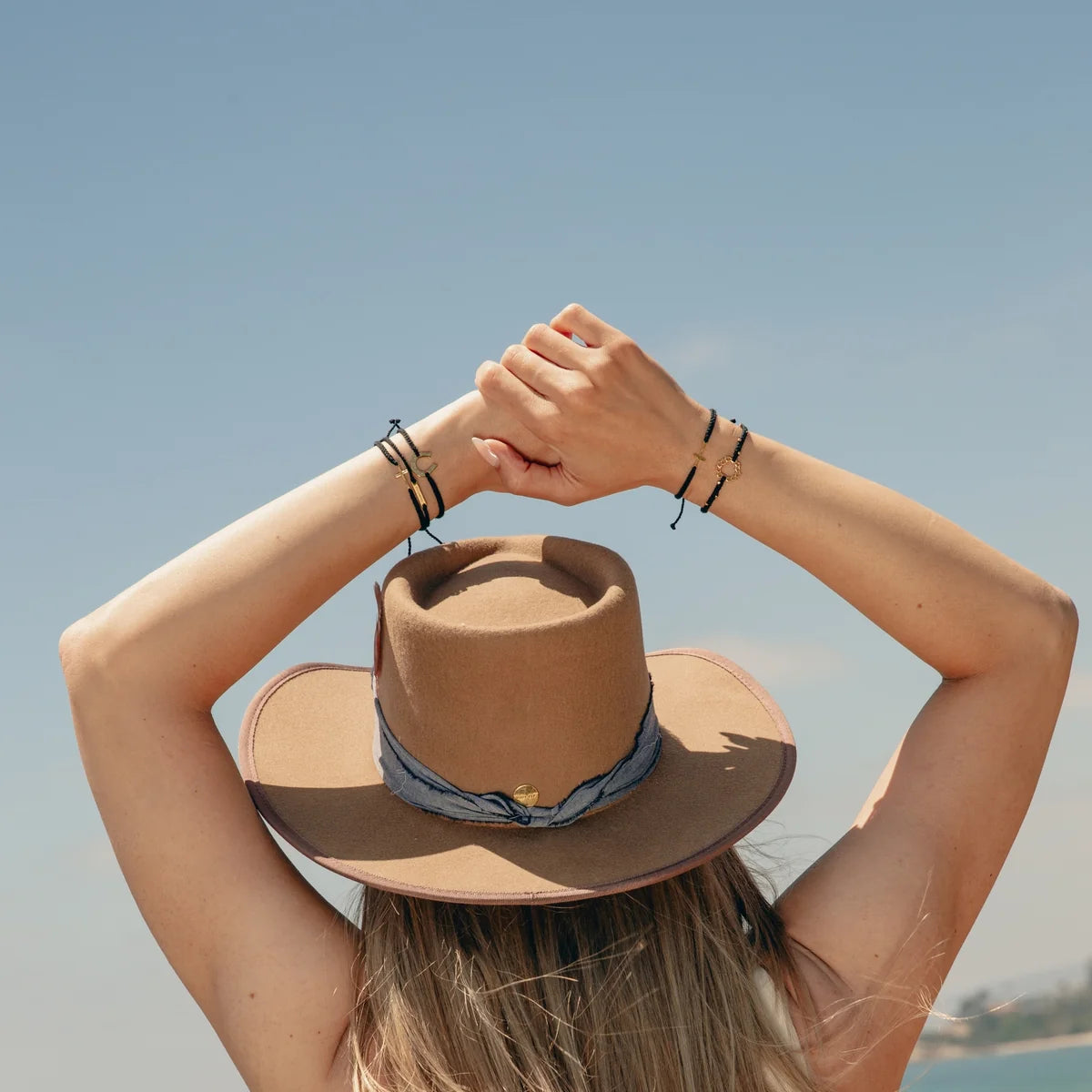 A woman viewed from behind wearing a tan wide-brim fedora hat with a blue patterned band and black bracelets on her raised arms, set against a clear blue sky.