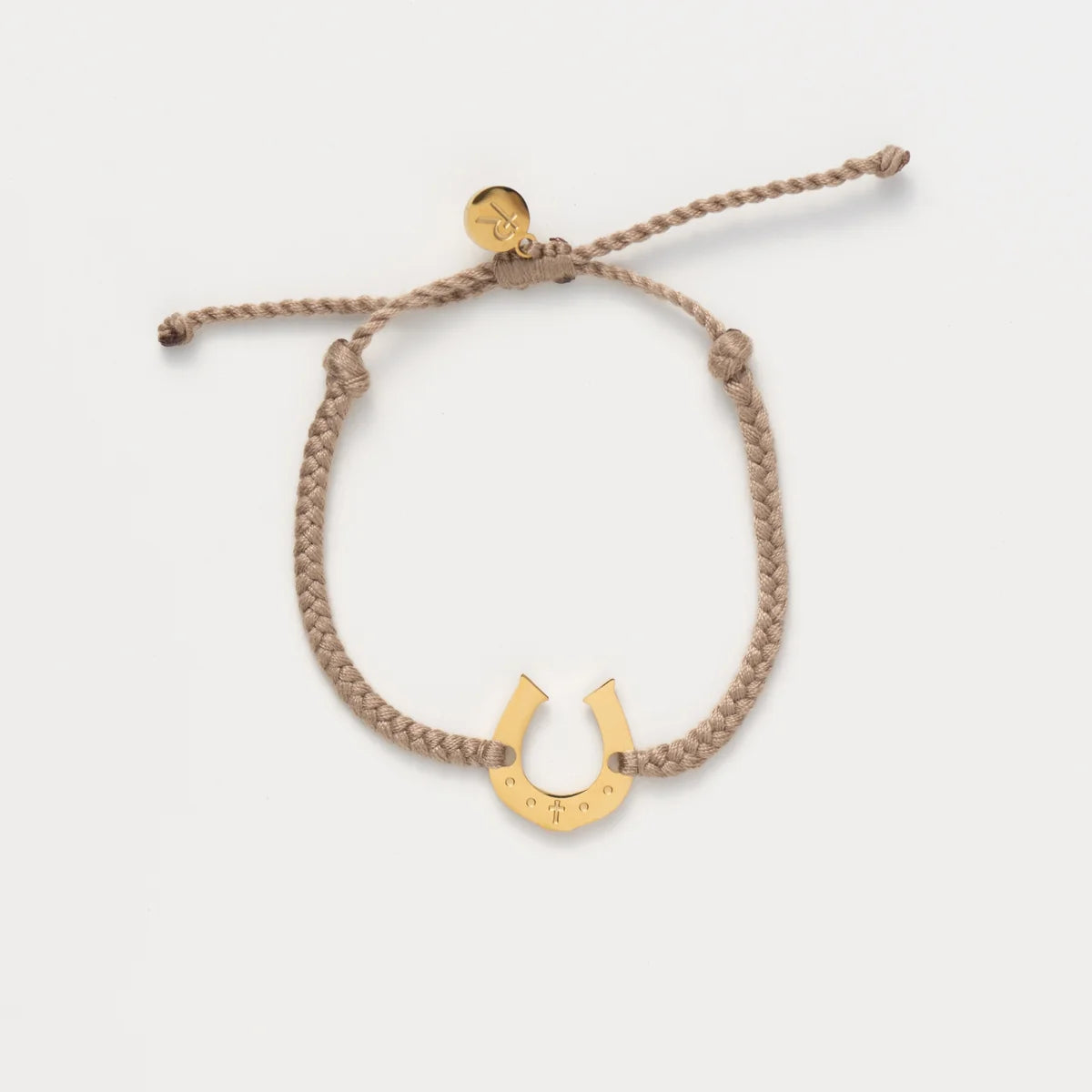 A braided tan rope bracelet with a gold horseshoe charm and a small round gold tag, displayed on a clean off-white background.
