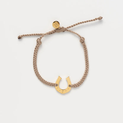 A braided tan rope bracelet with a gold horseshoe charm and a small round gold tag, displayed on a clean off-white background.