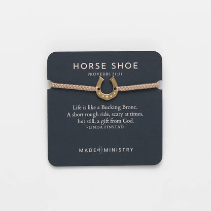 A horse shoe charm bracelet with a gold-colored metal horseshoe pendant and a beige braided cord, presented on a dark gray card that features a Bible verse and the brand name Made Ministry.