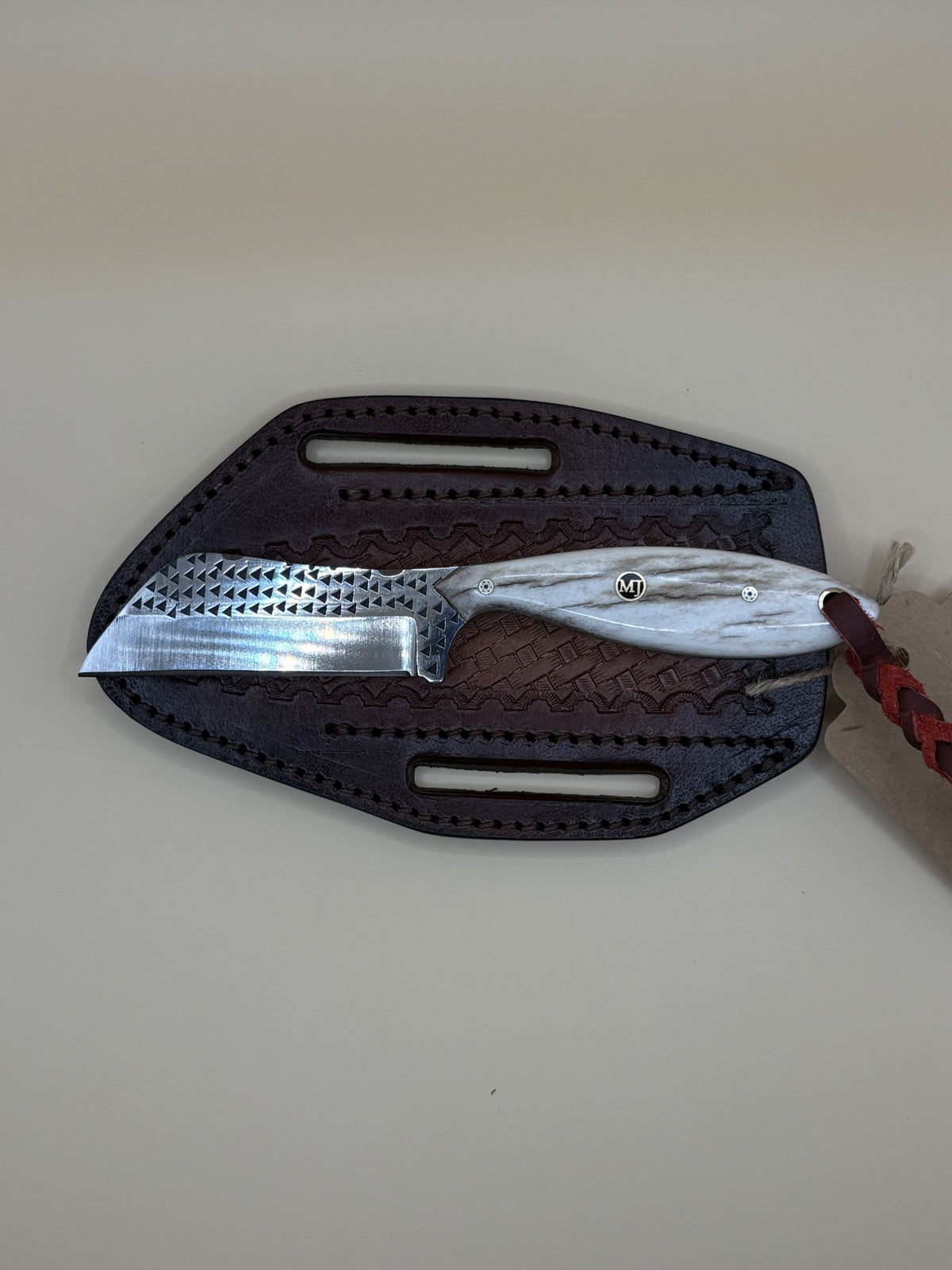 A fixed-blade knife with a hammered silver blade and a white marbled handle, resting on a dark brown textured leather sheath against a plain beige background.